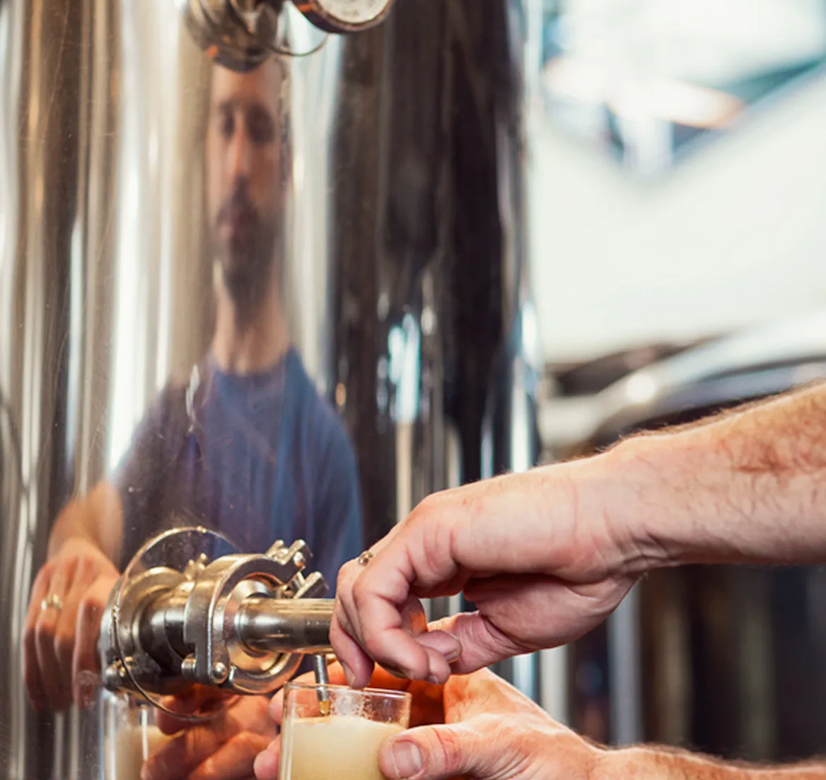 Close-up of a person filling a glass with beer from a metal tap in a brewery, with the person's reflection on the shiny tank.