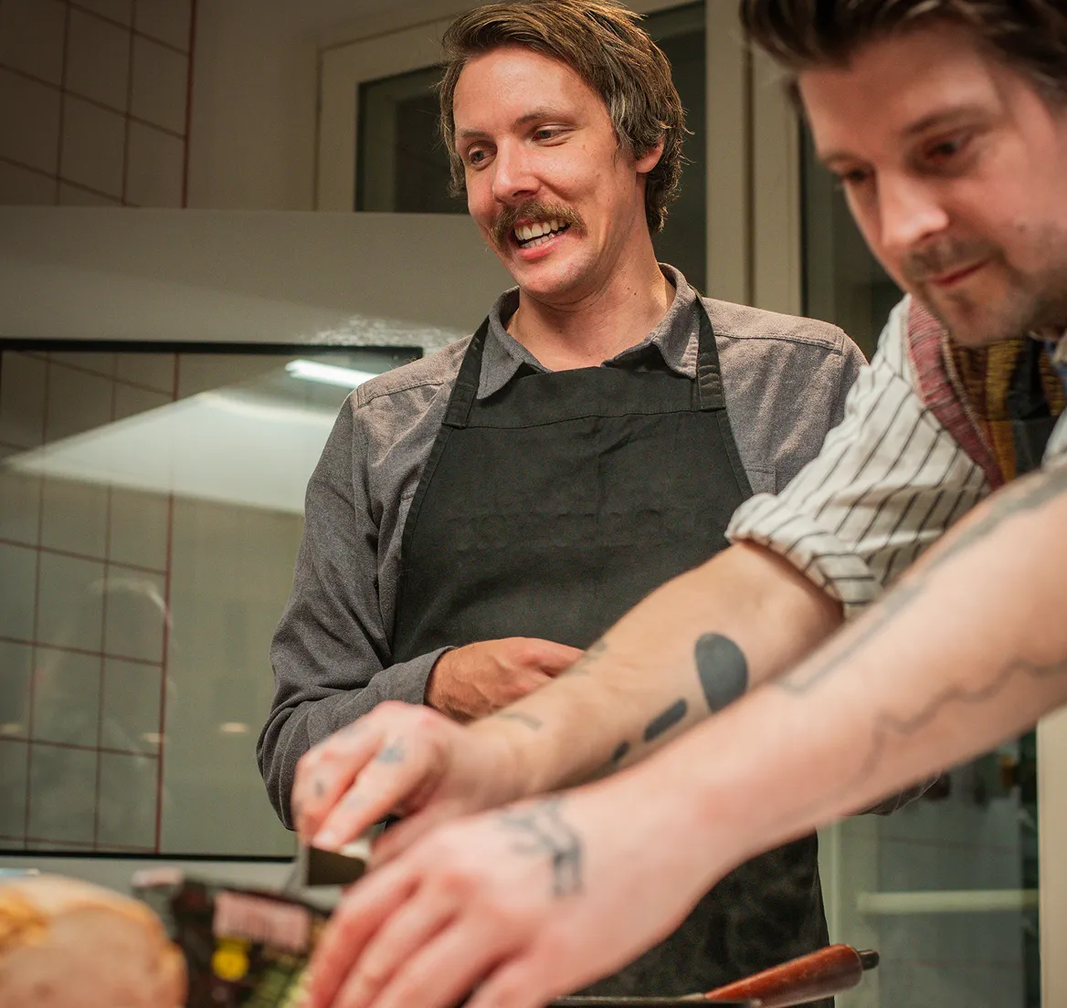 Two men preparing food in a kitchen, one wearing a black apron and smiling, the other focused on the task.