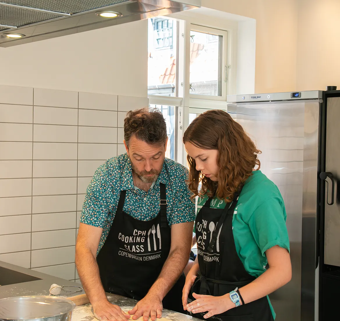 Man and woman wearing black aprons preparing dough together in a kitchen with white tiled wall and stainless steel refrigerator.
