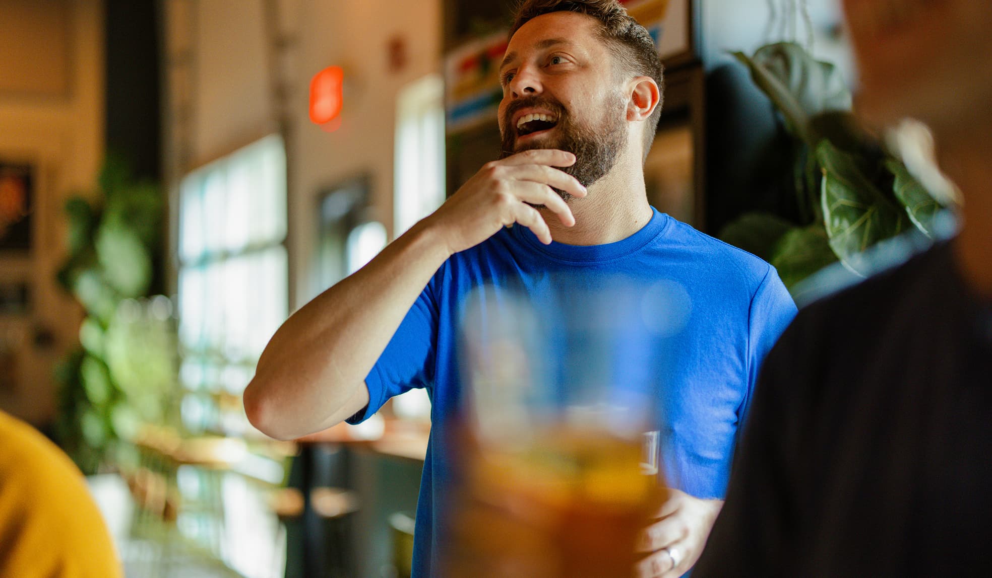 Bearded man wearing a blue shirt smiling and touching his chin with a blurred glass in the foreground.