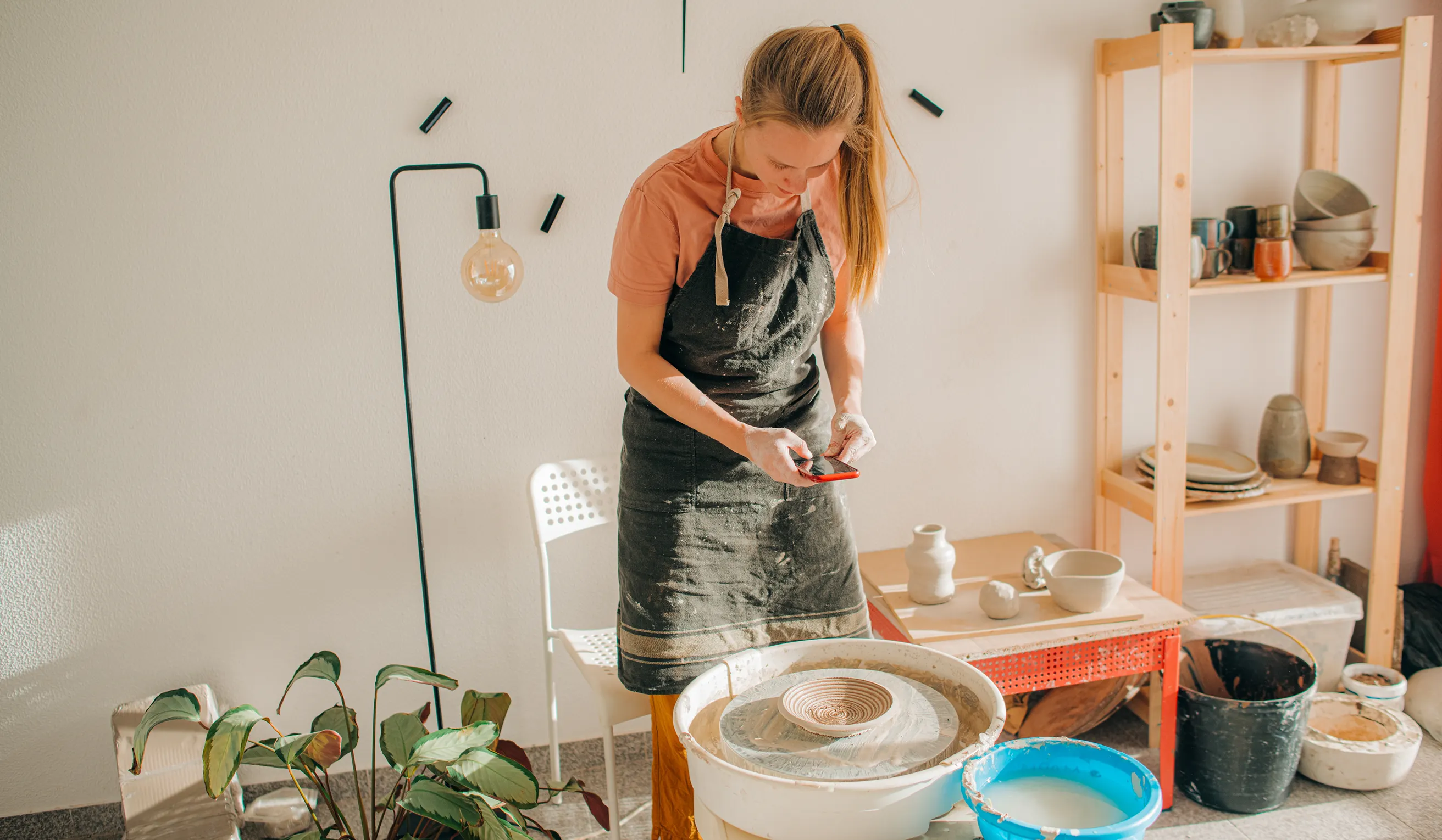 Woman in a black apron working with a pottery wheel in a sunlit studio.