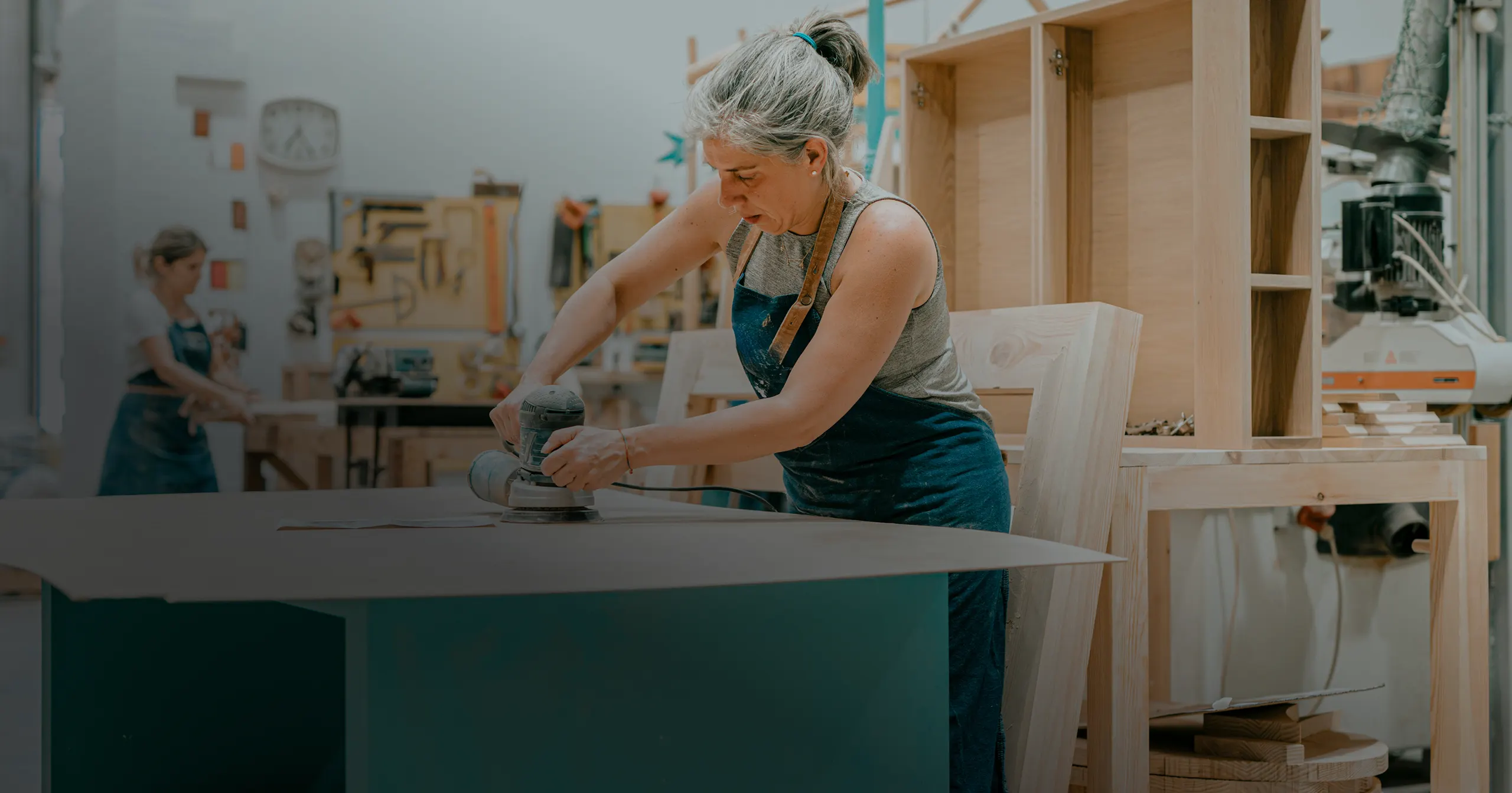 Woman in a workshop using a power sander on a large wooden surface.