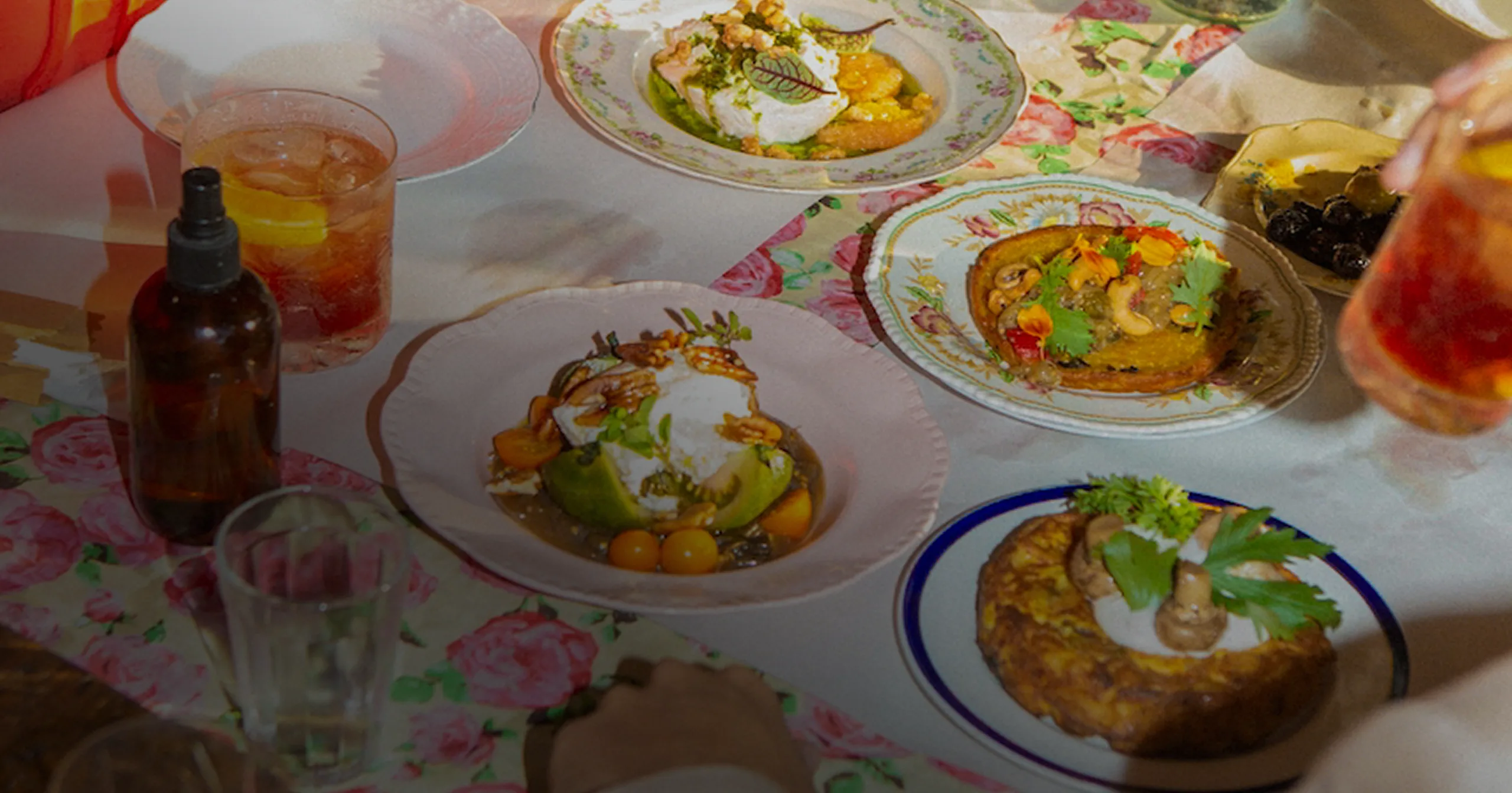 Table set with various gourmet dishes and drinks, including plates with salads, garnished appetizers, and cocktails on floral tablecloth.