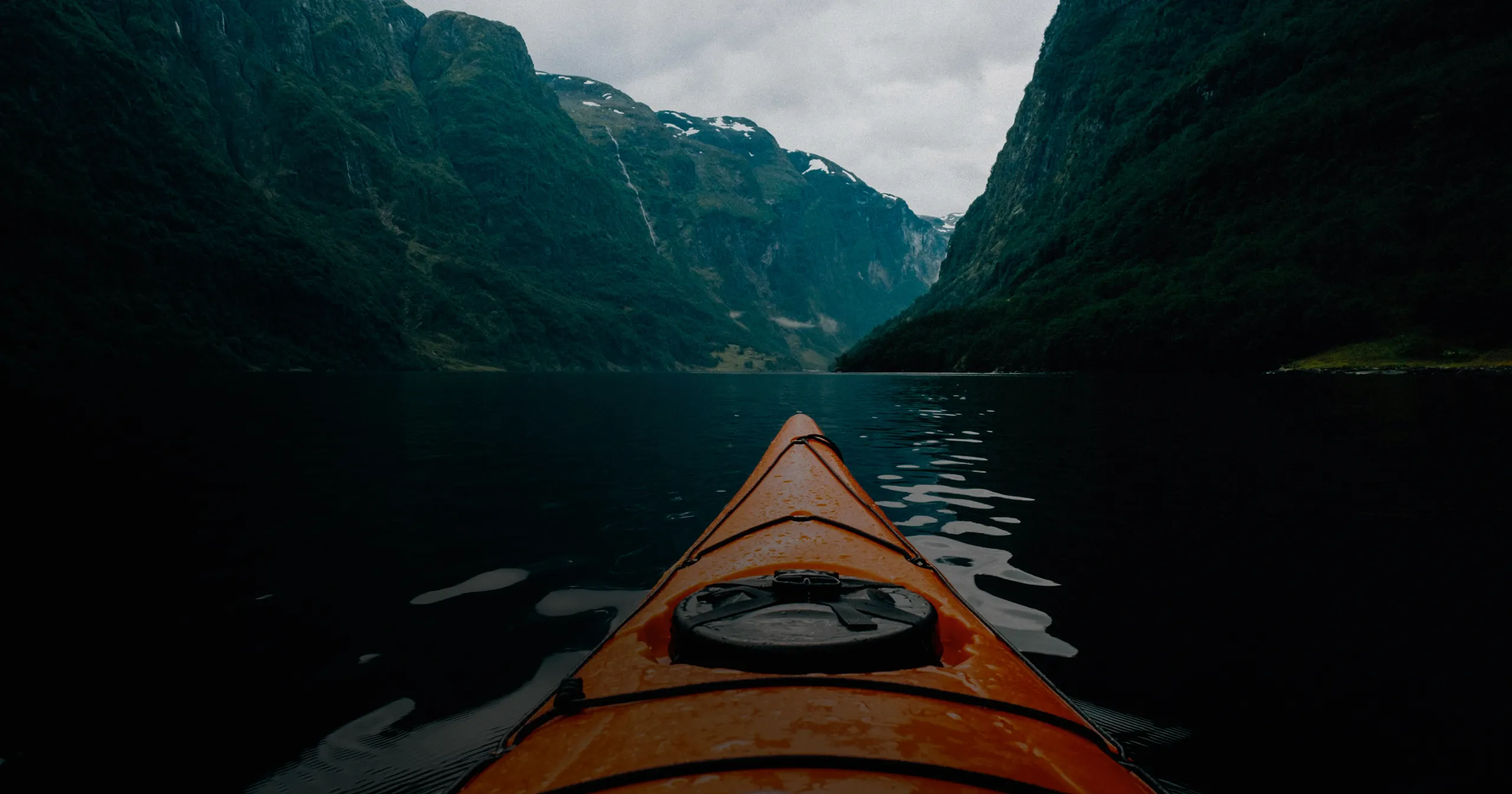 View from a kayak on calm water surrounded by steep, green mountains under a cloudy sky.