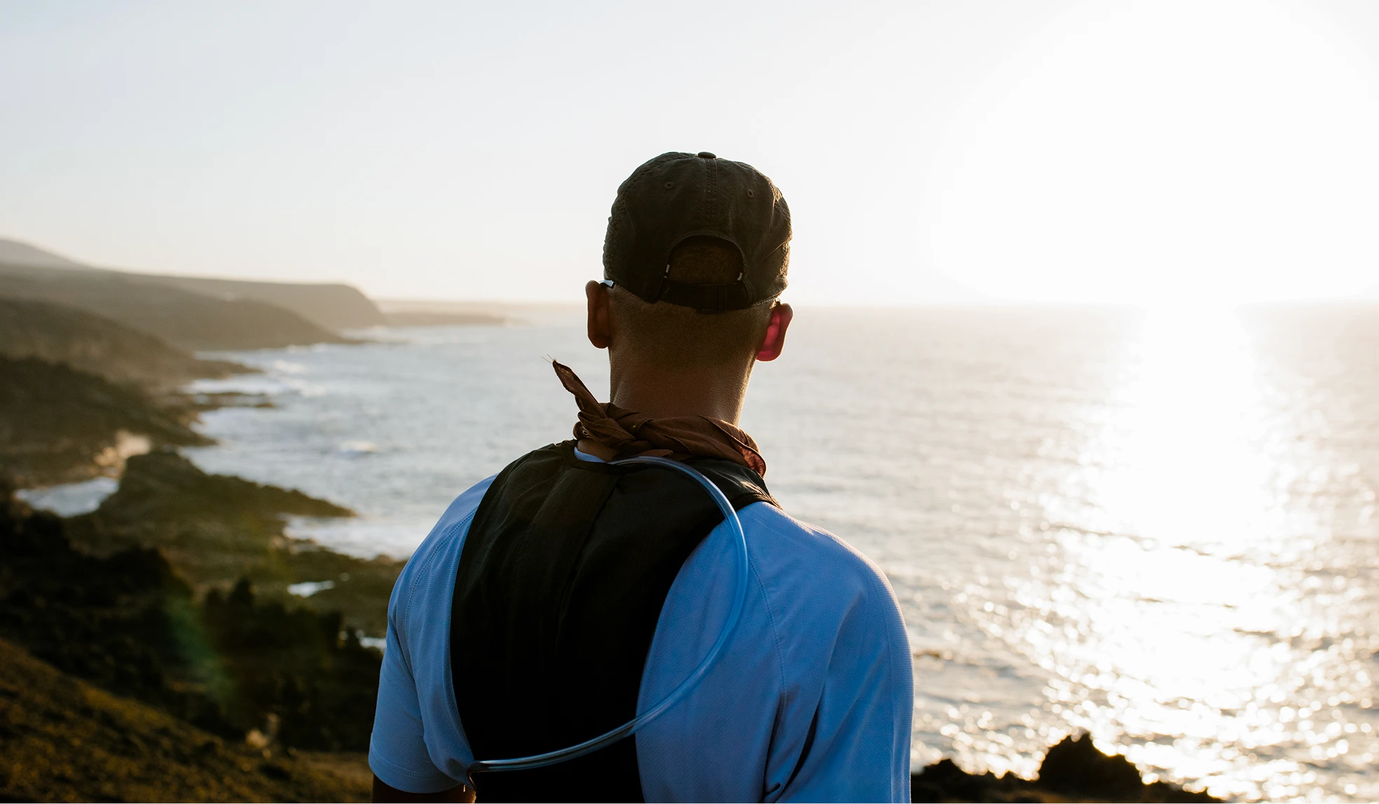 Person wearing a cap and hydration pack looking out over a rocky coastline at sunset.