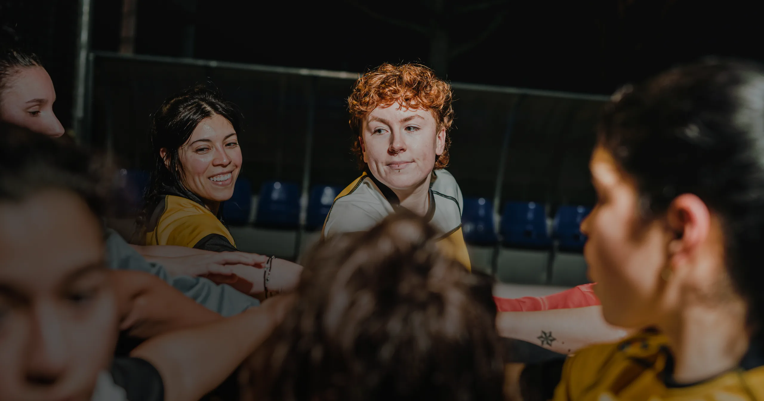 A diverse group of women athletes in sports jerseys huddle together with hands joined in a gesture of team unity on a dark sports field.
