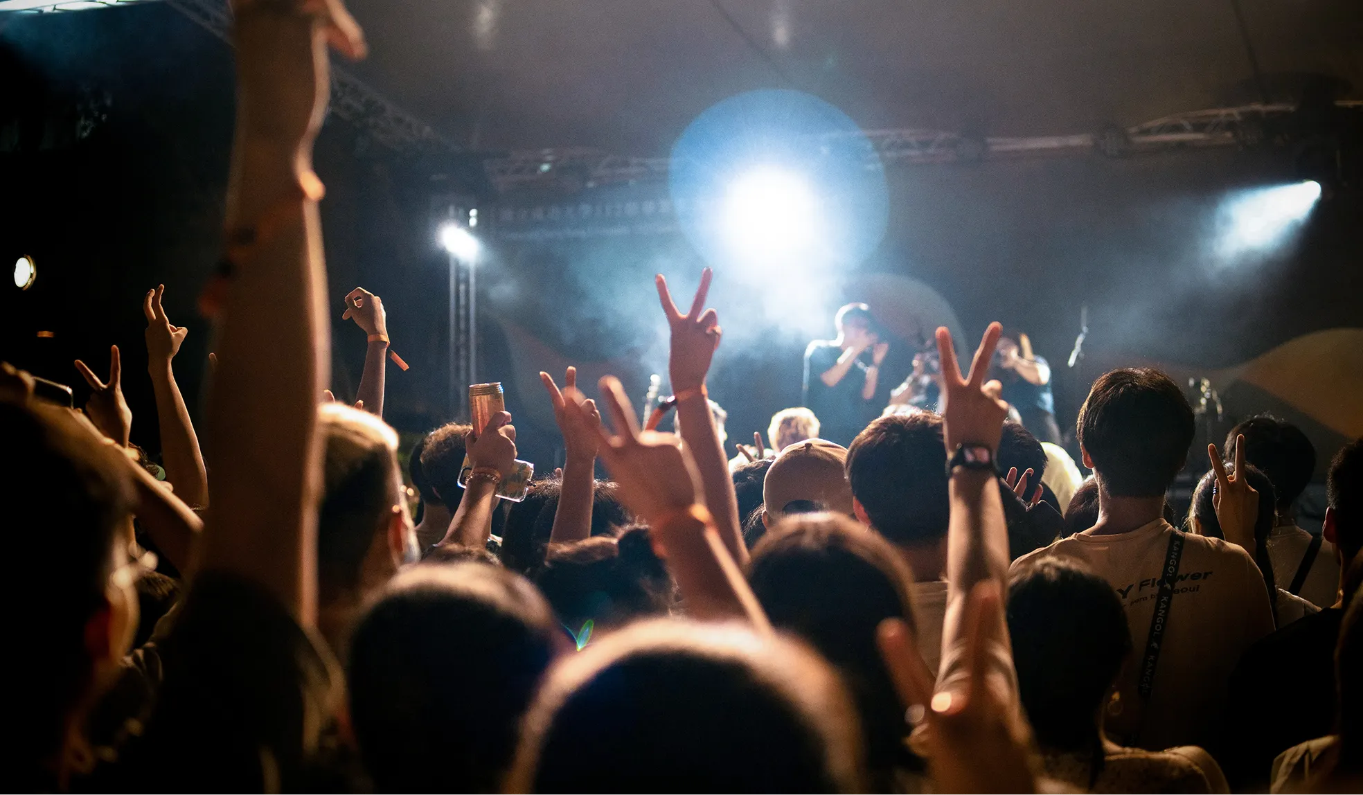Crowd at a concert raising hands and making peace signs toward a brightly lit stage with performers.