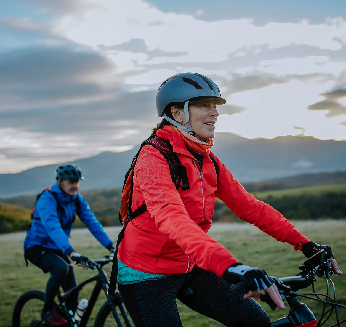 Two cyclists wearing helmets and jackets riding mountain bikes outdoors with hills and cloudy sky in the background.