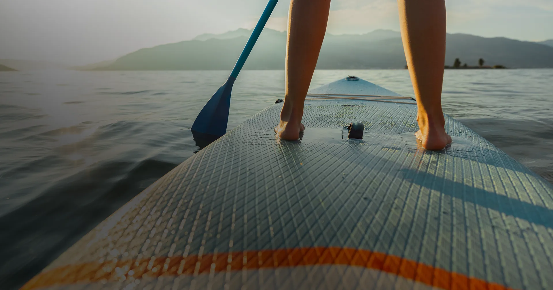 Close-up of a person's bare feet on a paddleboard in calm water with mountains in the background.
