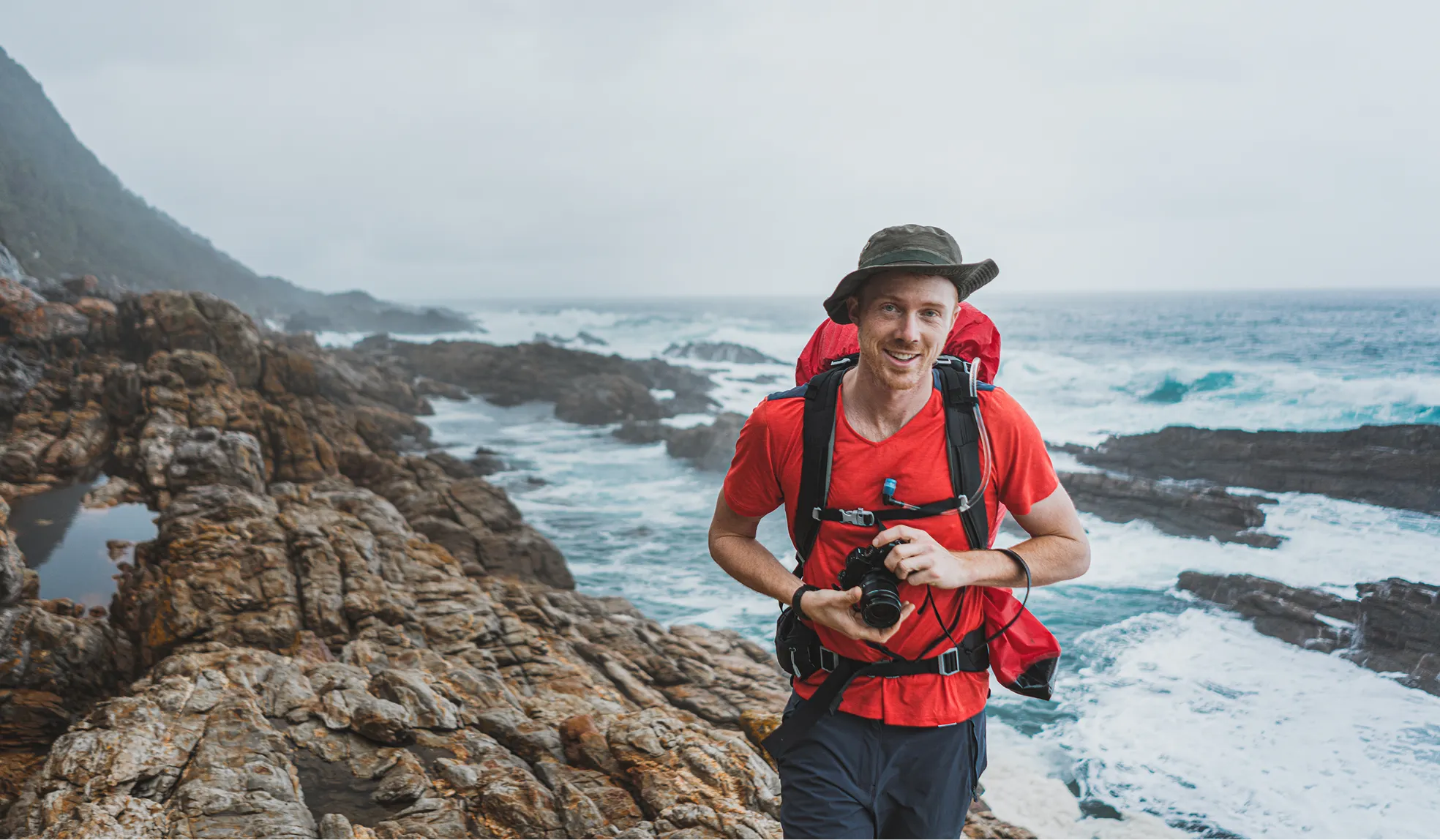 Smiling man hiking on rocky coastline with a camera and red backpack near ocean waves.