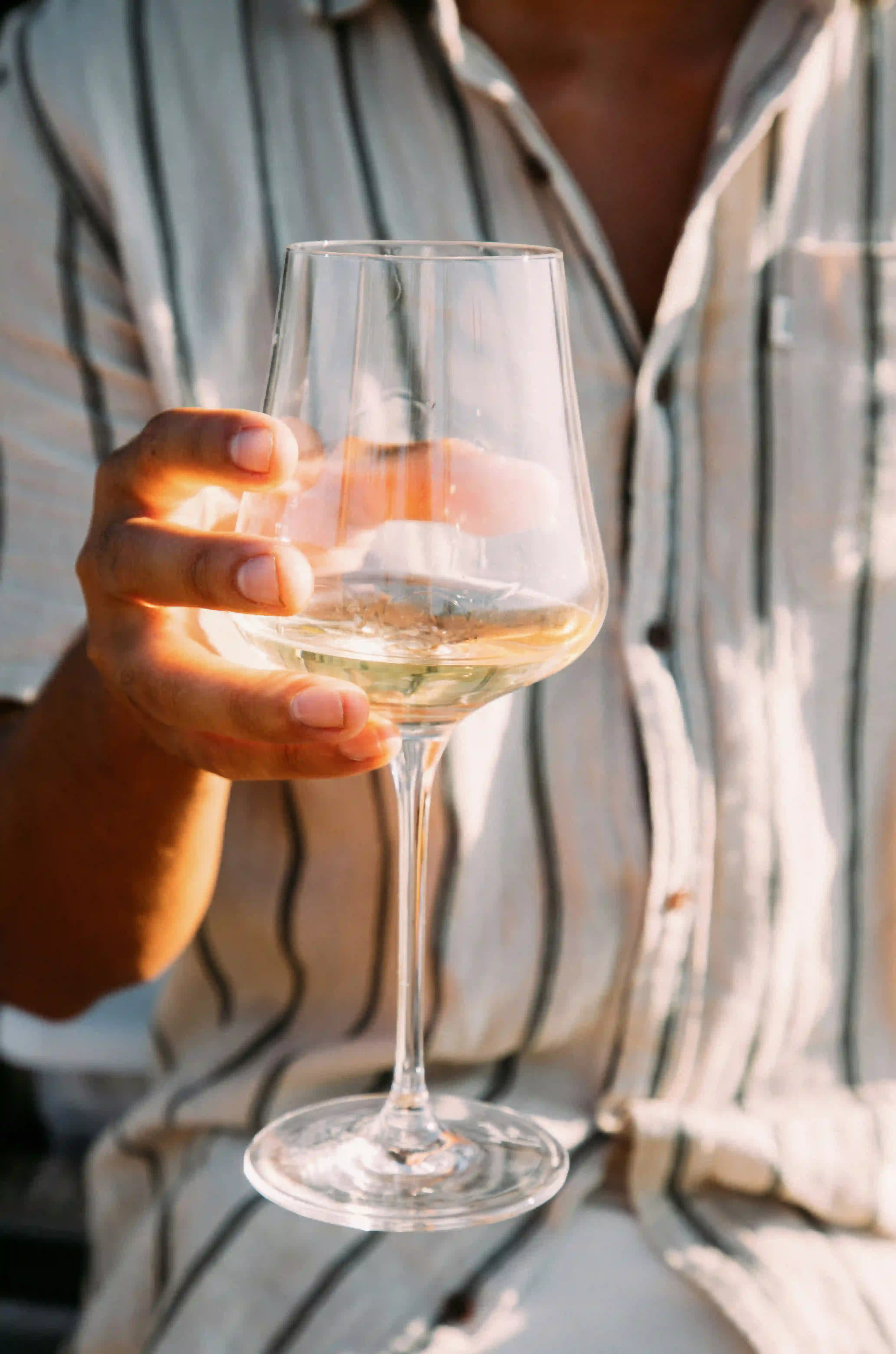 Close-up of a person holding a wine glass with white wine, wearing a light shirt with vertical stripes.