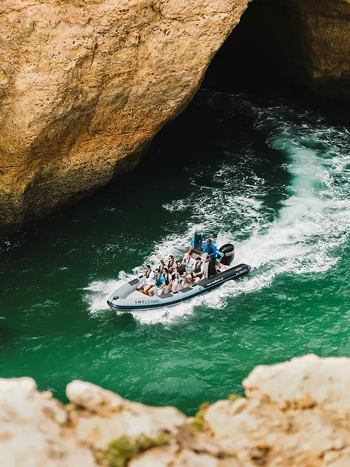 Small motorboat with passengers wearing life jackets cruising through green water near rocky cliffs and a cave entrance.