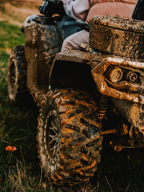 Close-up of a muddy all-terrain vehicle tire and front suspension on grass.