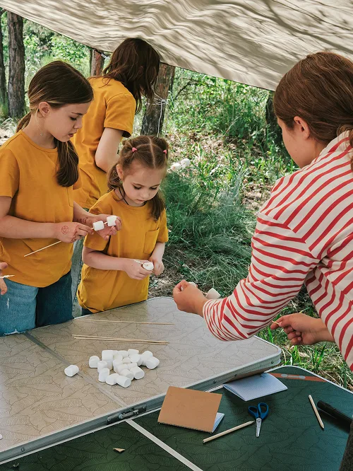 Three children in yellow shirts preparing marshmallows on skewers at a table outdoors with an adult in a red and white striped shirt assisting them.