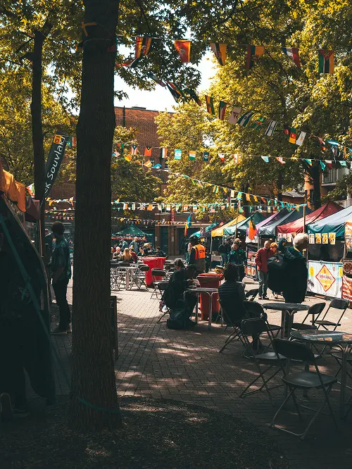 Outdoor market with people sitting and walking among colorful tents and international flags strung above in a tree-lined area.