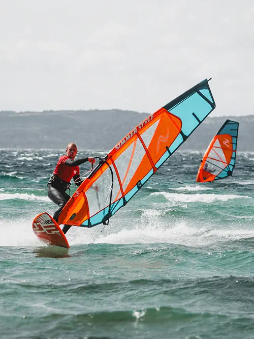 Person windsurfing on choppy ocean water with an orange and blue sail, another windsurfer visible in background.