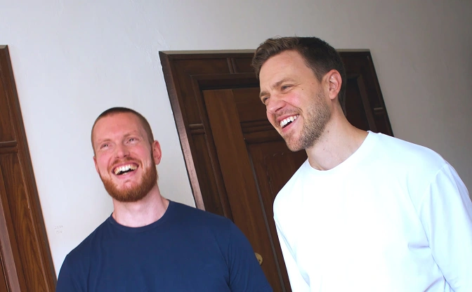 Two men smiling and laughing indoors in front of wooden doors on a white wall.