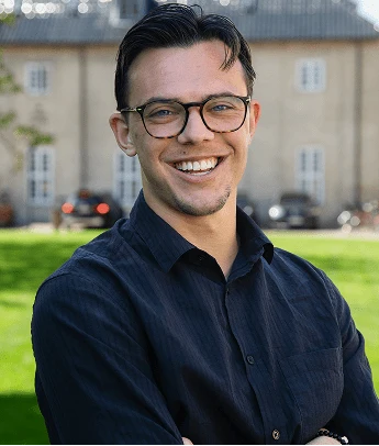 Young man with glasses smiling outdoors in front of a building with green grass.