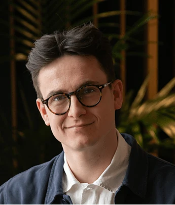 Man with glasses smiling, wearing a white shirt and dark jacket in a dimly lit indoor setting.