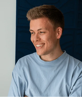 Young man with short brown hair wearing a light blue shirt smiling and looking to the side.