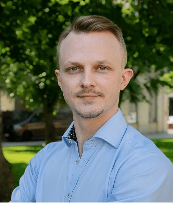 Man with short light brown hair and blue eyes in a blue shirt standing outdoors with trees in background.