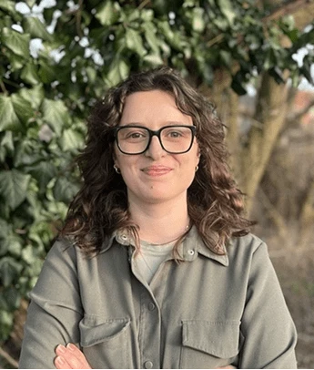 Smiling woman with curly hair and glasses wearing a gray button-up shirt outdoors.