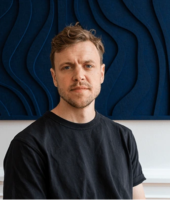 Man with short brown hair and beard wearing a black t-shirt against a dark blue patterned wall.