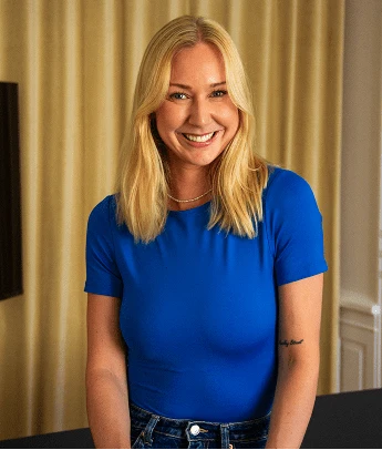 Smiling woman with long blonde hair wearing a blue shirt and jeans standing indoors.