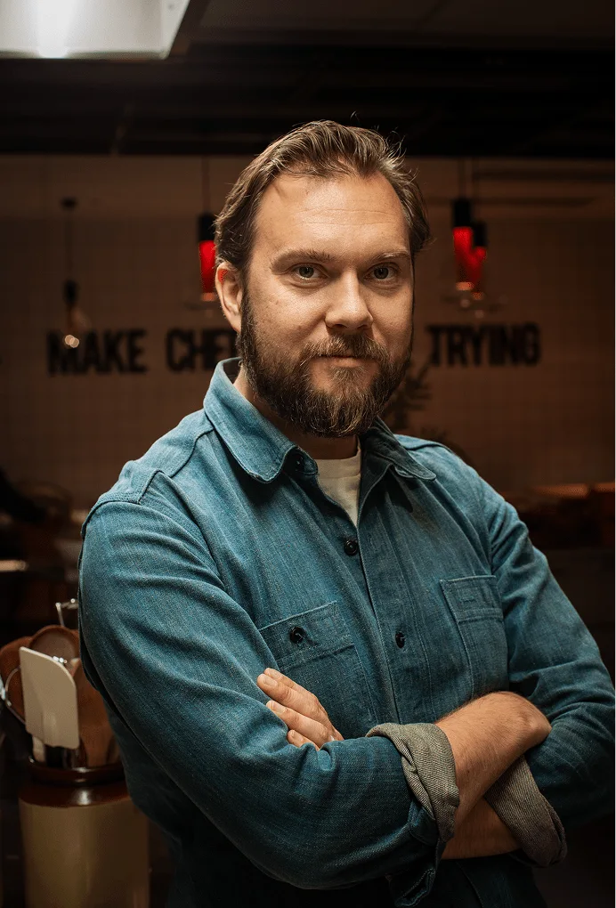 Man with beard and denim shirt stands with arms crossed in a dimly lit room with red lights.
