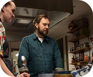 Two men focused on cooking in a kitchen with shelves holding spices and dishes.