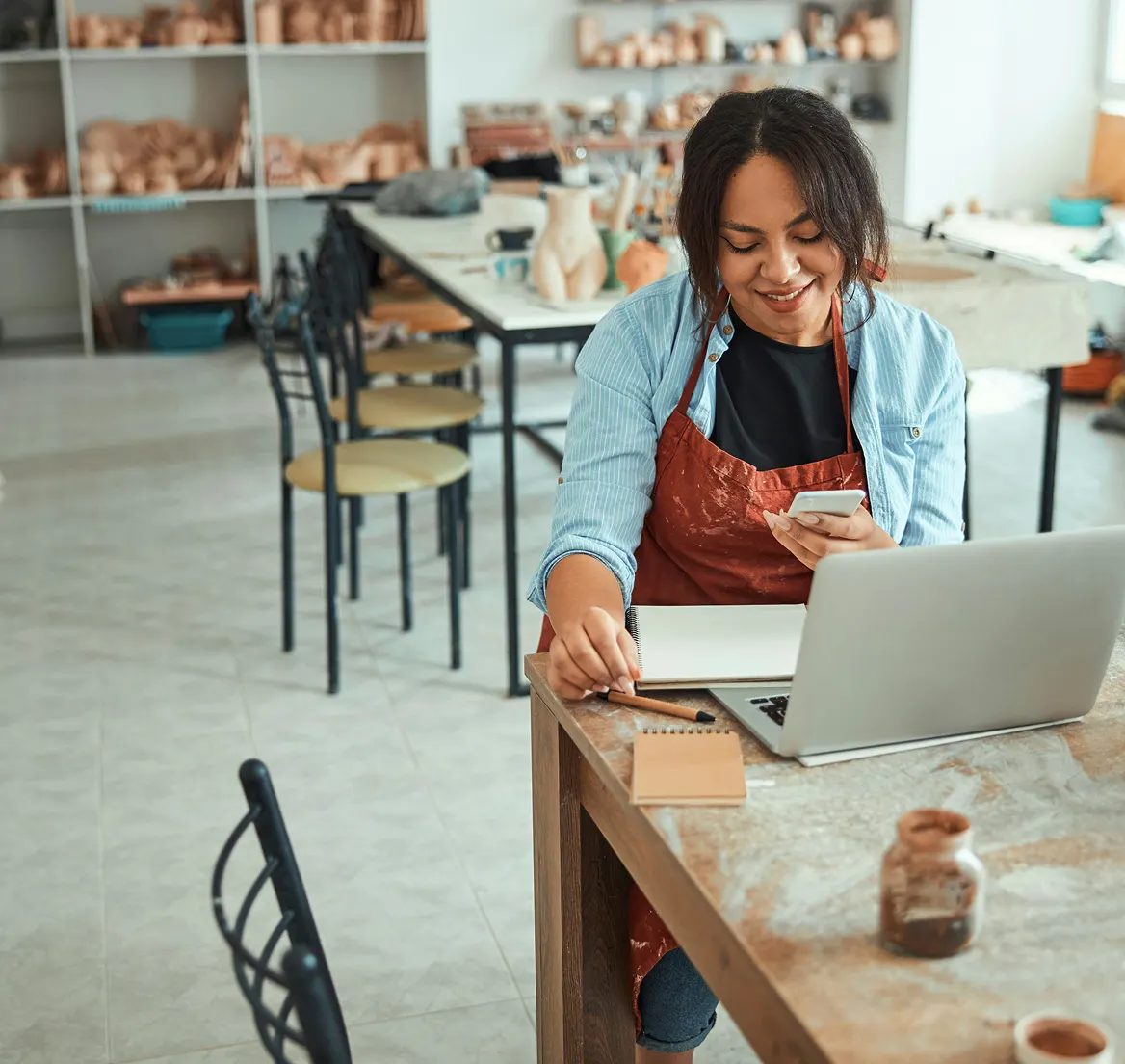 Smiling woman in artist apron using phone and laptop at a pottery studio table.