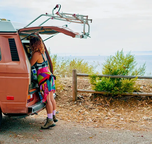 Person with curly hair wearing a colorful skirt and hiking boots leaning into the back of a rust-colored van.