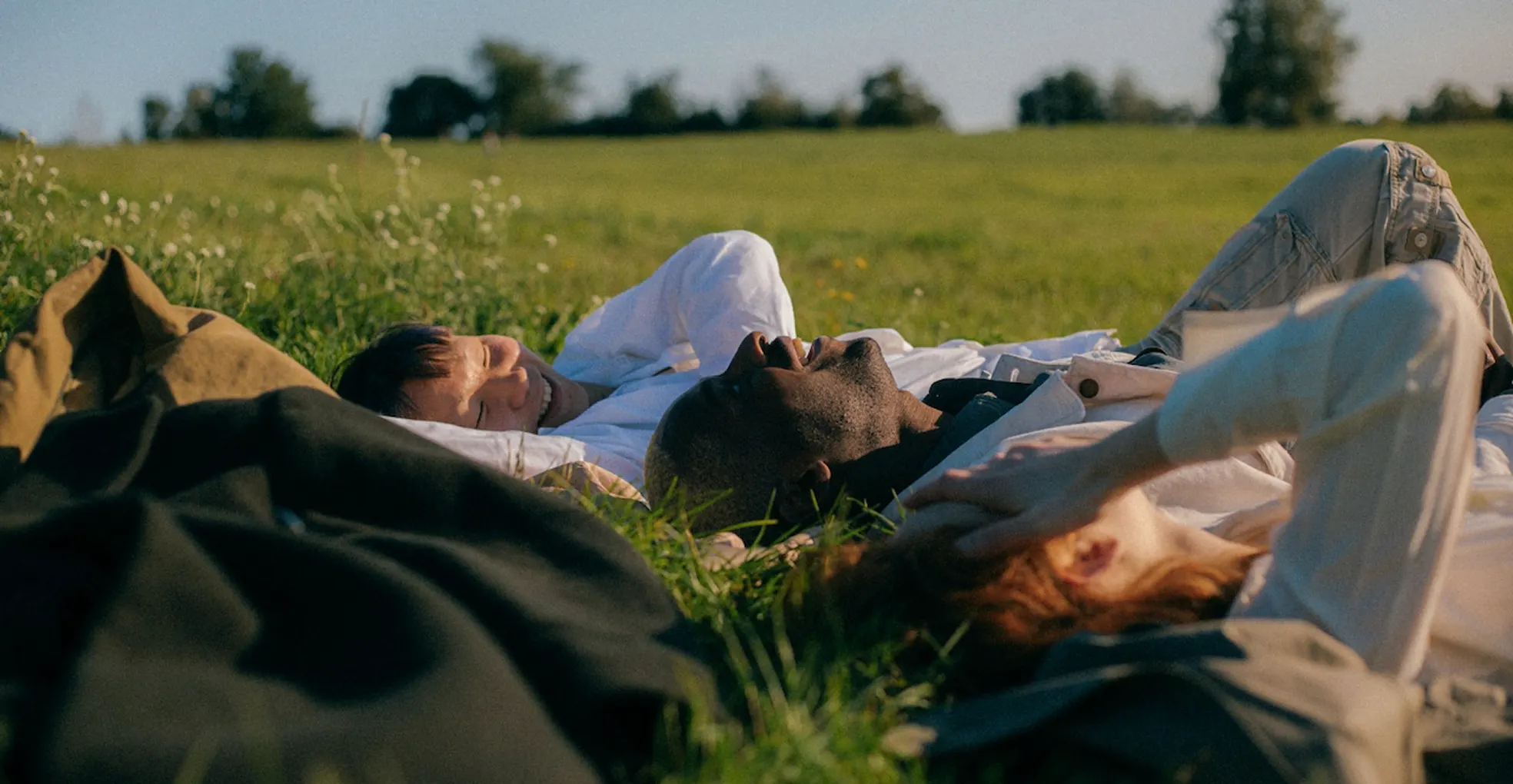 Three people lying on grass outdoors on a sunny day, relaxing and smiling in a green field.