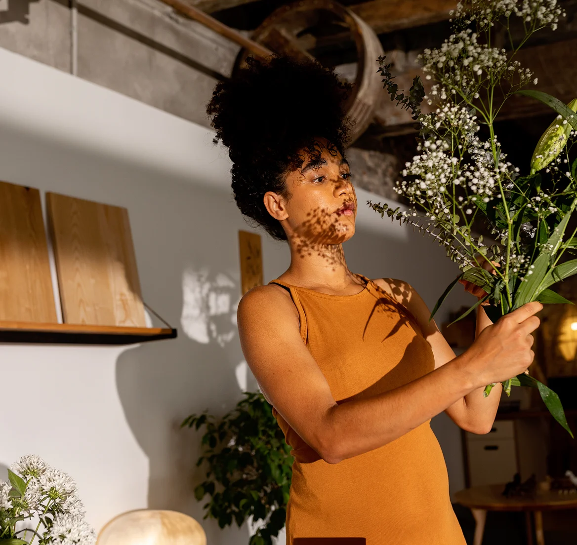 Woman in orange sleeveless dress arranging white flowers and green leaves indoors with shadows on her face.