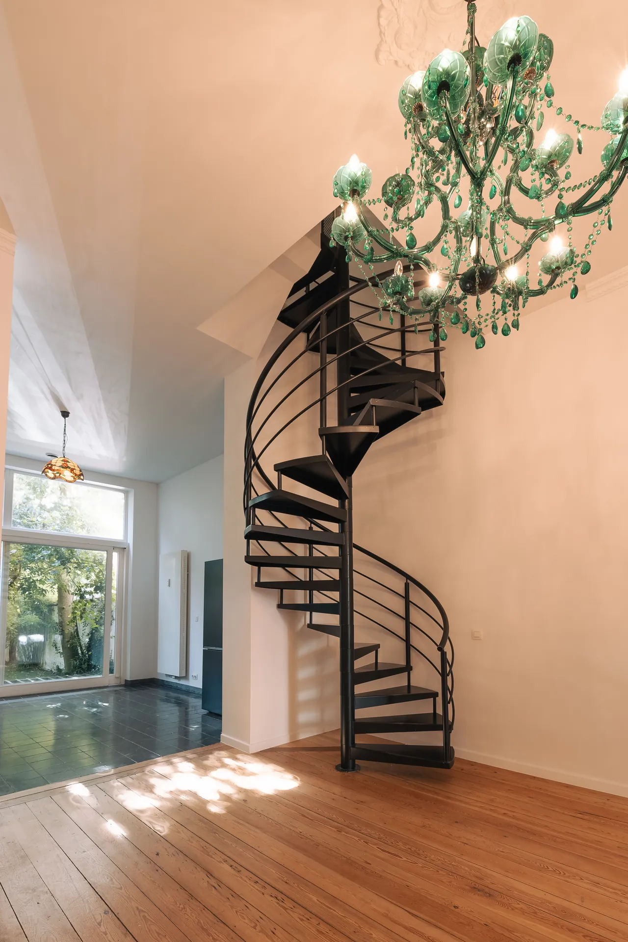 Escalier en colimaçon métallique sous un lustre en verre, séjour baigné de lumière — maison rénovée à Saint-Gilles, Bruxelles