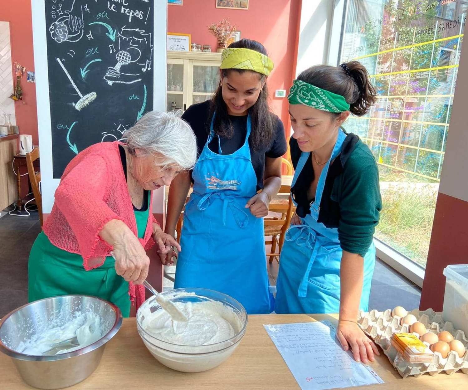 Trois femmes, dont une personne âgée, cuisinent ensemble dans une cuisine conviviale. Elles portent des tabliers bleus et sourient autour d’un grand saladier de pâte, symbolisant le partage intergénérationnel et la transmission culinaire.