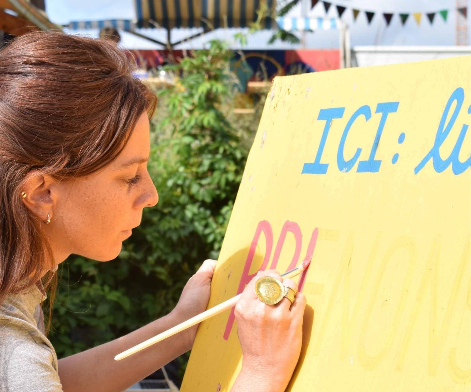Jeune femme concentrée en train de peindre à la main un panneau coloré portant le mot “ICI”, lors d’un atelier en plein air, illustrant un engagement créatif et local.