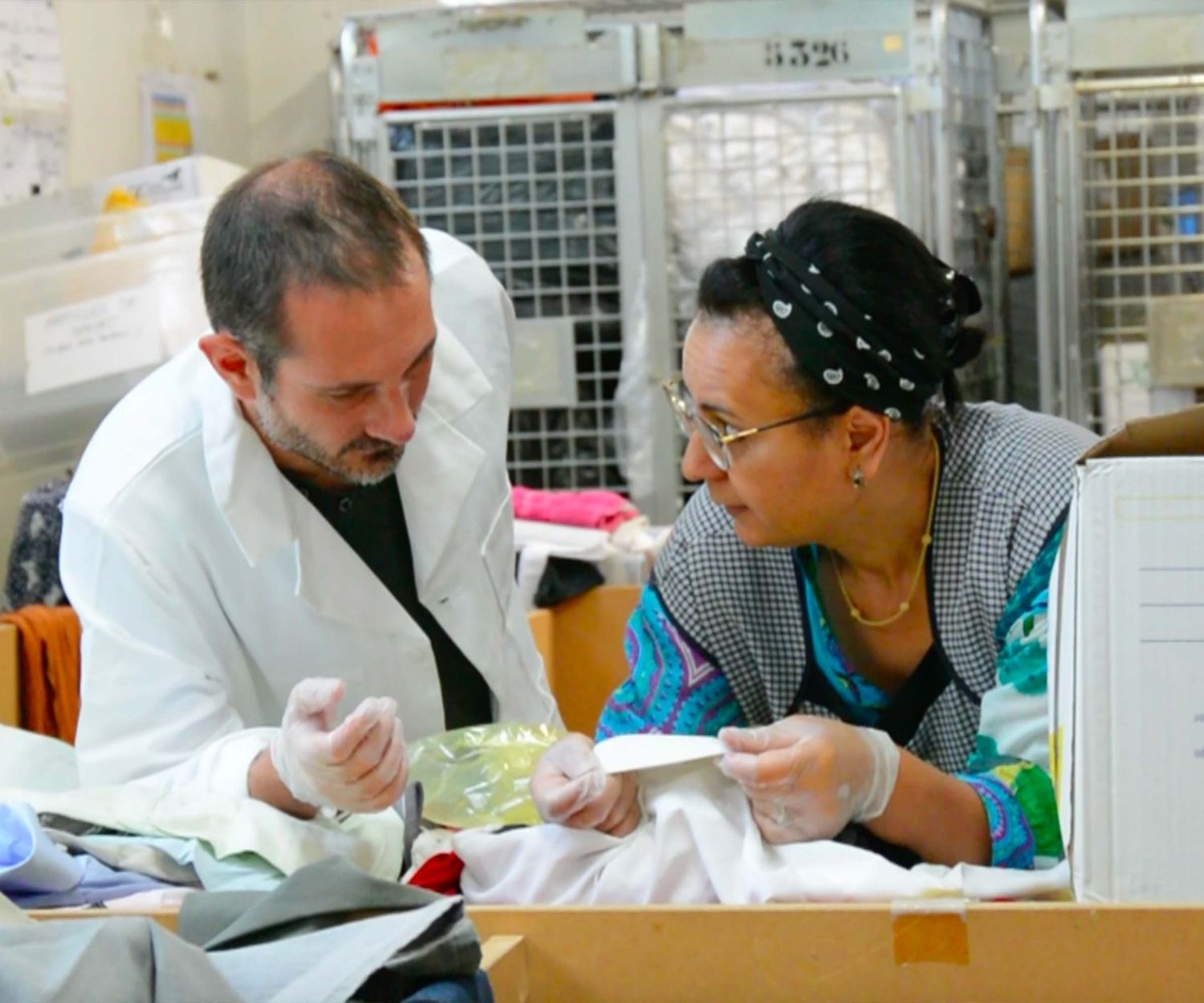Deux personnes portent des gants et trient des vêtements dans un atelier de recyclage textile, échangeant de manière attentive. Une scène de travail solidaire et circulaire en milieu logistique.