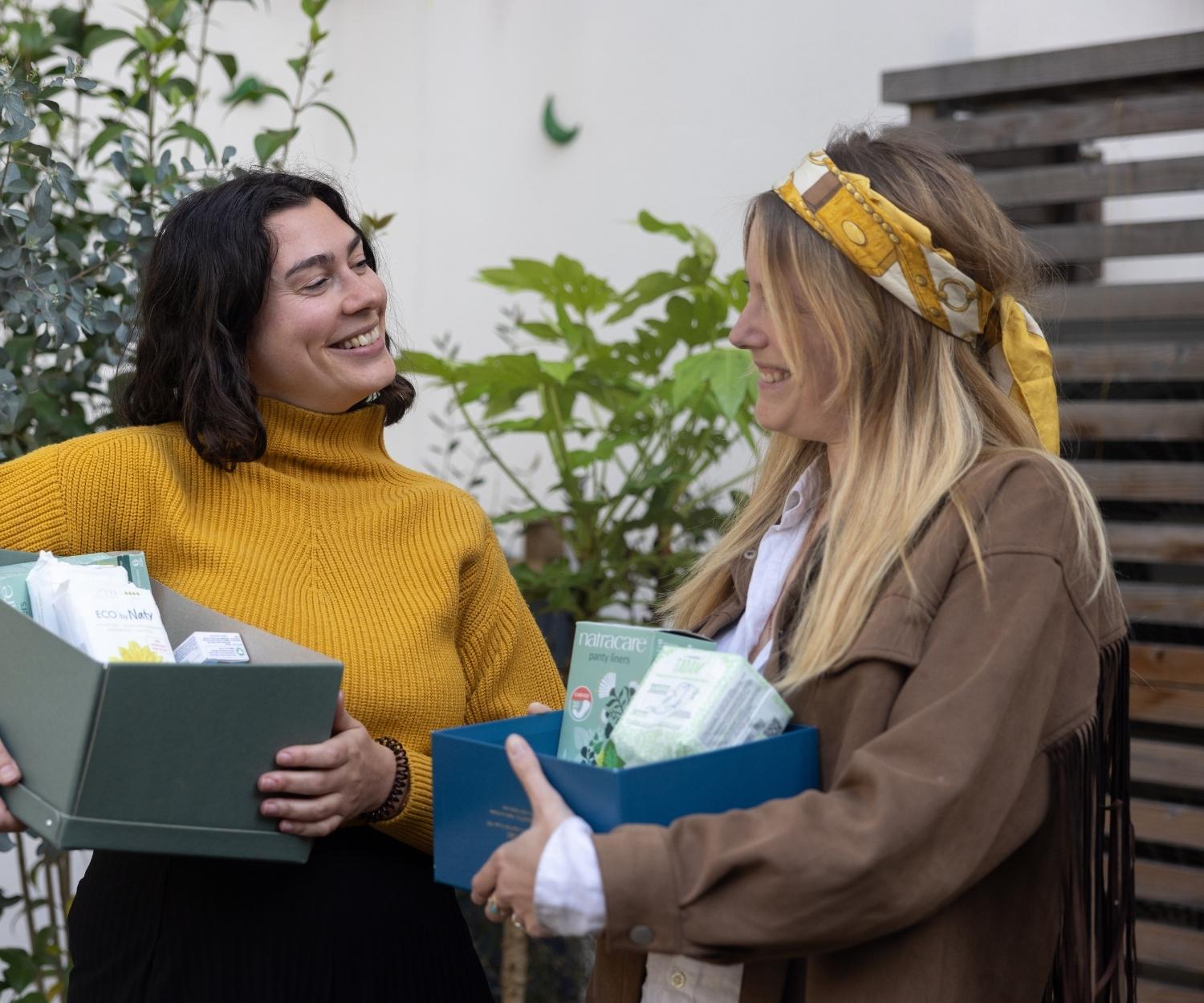 Deux femmes souriantes s’échangent des boîtes remplies de produits d’hygiène, dans un cadre extérieur verdoyant. Une scène de solidarité et de partage autour de la précarité menstruelle.