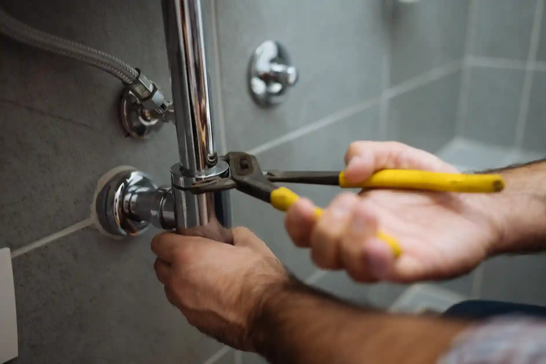  Hands using pliers to install chrome plumbing pipes against a tiled wall.