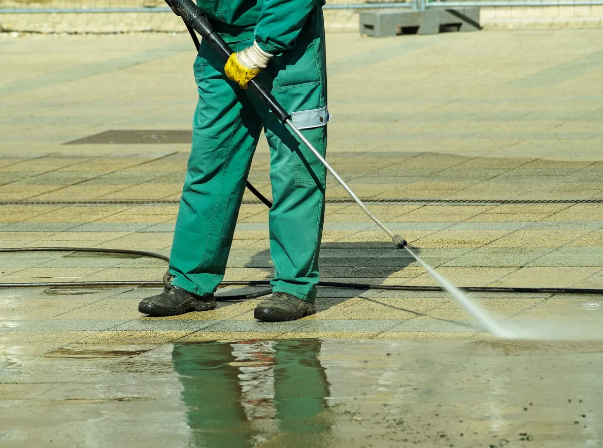 A person in green overalls and yellow gloves uses a high-pressure water jet to clean a paved surface.