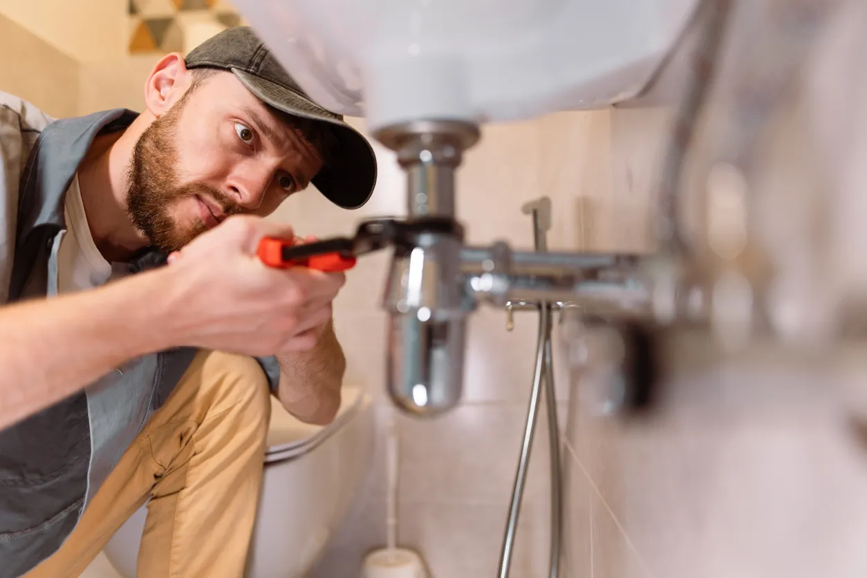 A man in a cap works on plumbing pipes under a sink in a bathroom, holding an adjustable wrench.