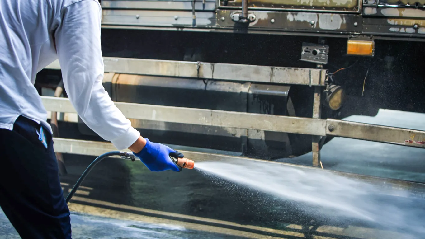 A person in a white long-sleeved shirt and blue gloves uses a high-pressure water jet to clean the side of a large dark vehicle or trailer.
