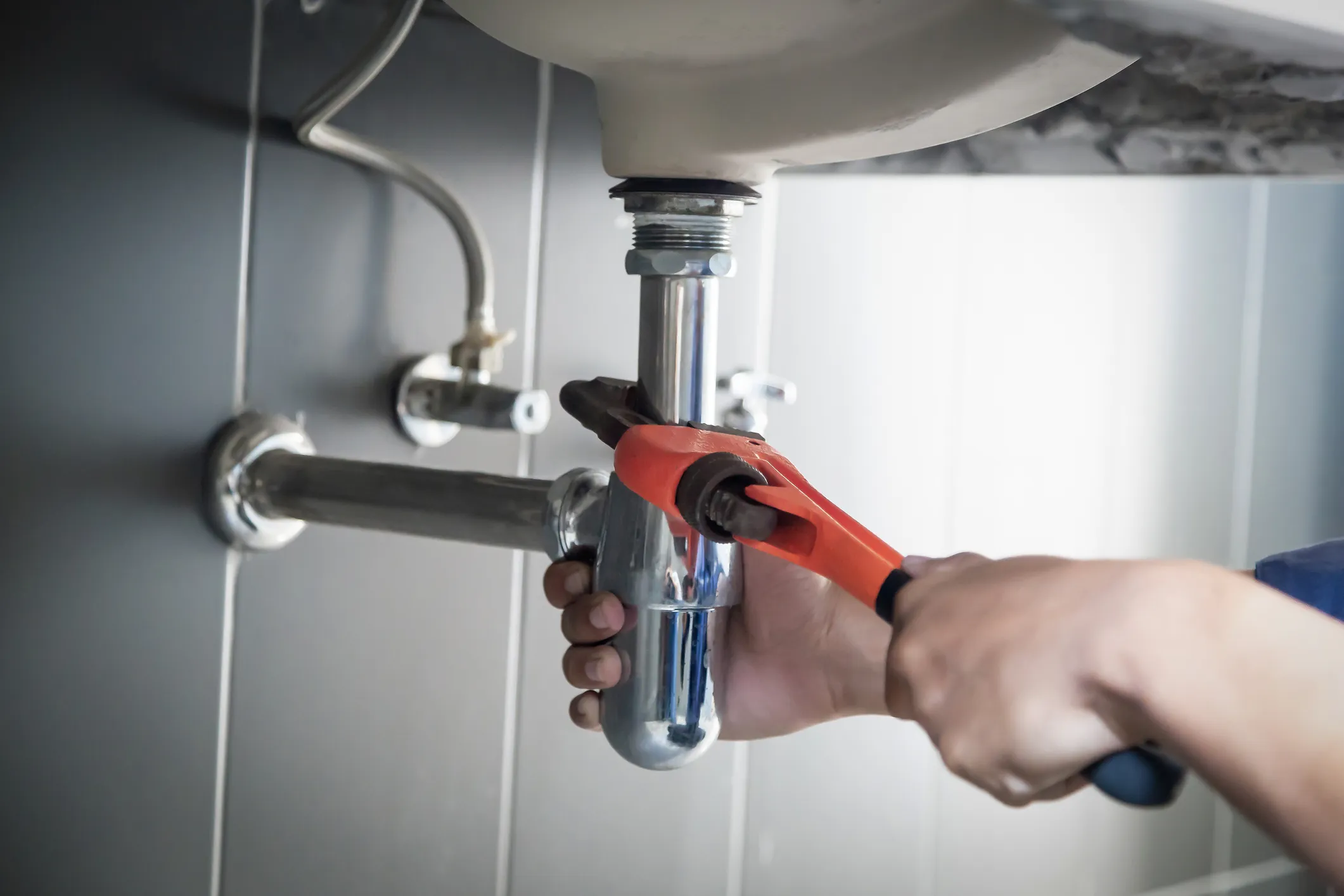 Hands using a wrench and holding a pink rag to work on chrome plumbing pipes under a white sink.