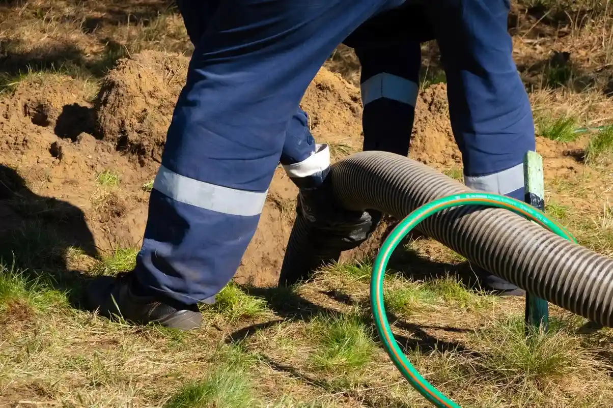 A person in blue overalls and gloves is using a large corrugated hose to pump out a septic tank, with only their lower body and the hose visible in the frame.