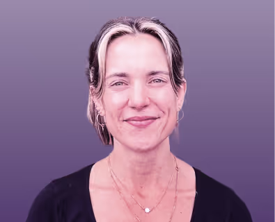 Smiling woman with light brown hair tied back, wearing hoop earrings and layered necklaces against a purple background.