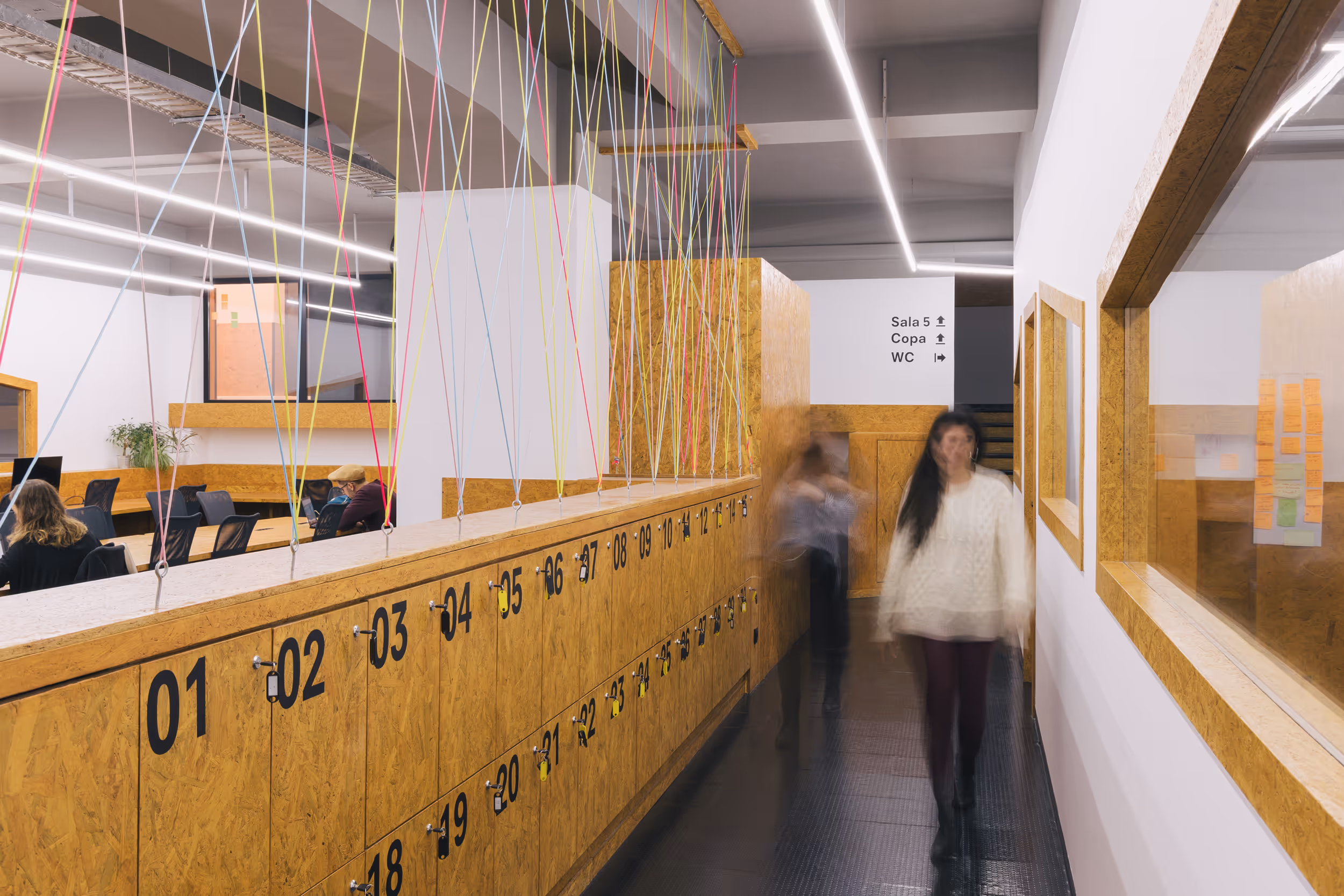 Modern office hallway with numbered wooden lockers on the left, colorful hanging strings above, and two blurred people walking past.