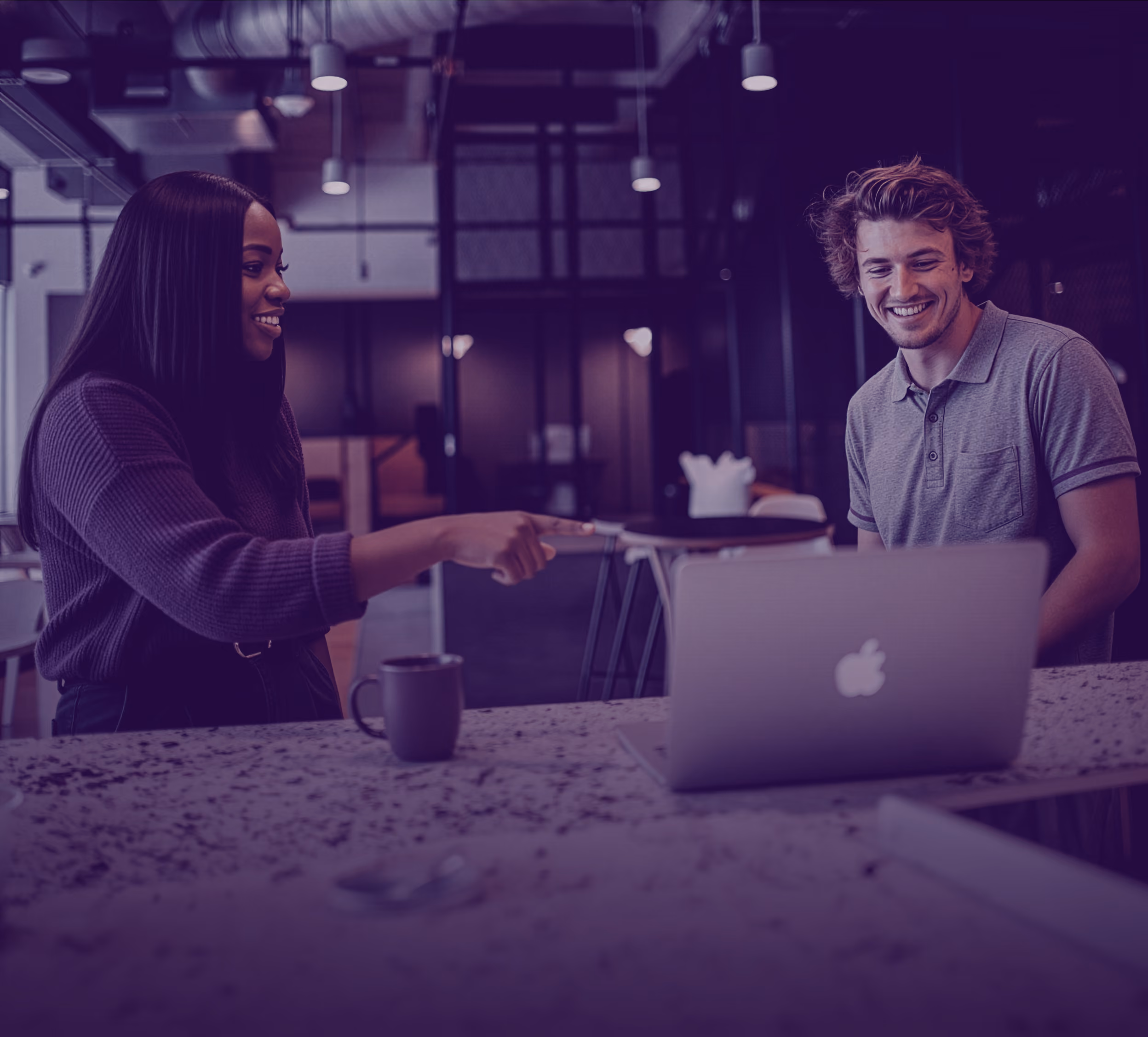 A woman pointing at a laptop screen while smiling, and a man sitting beside her also smiling in a modern office setting.