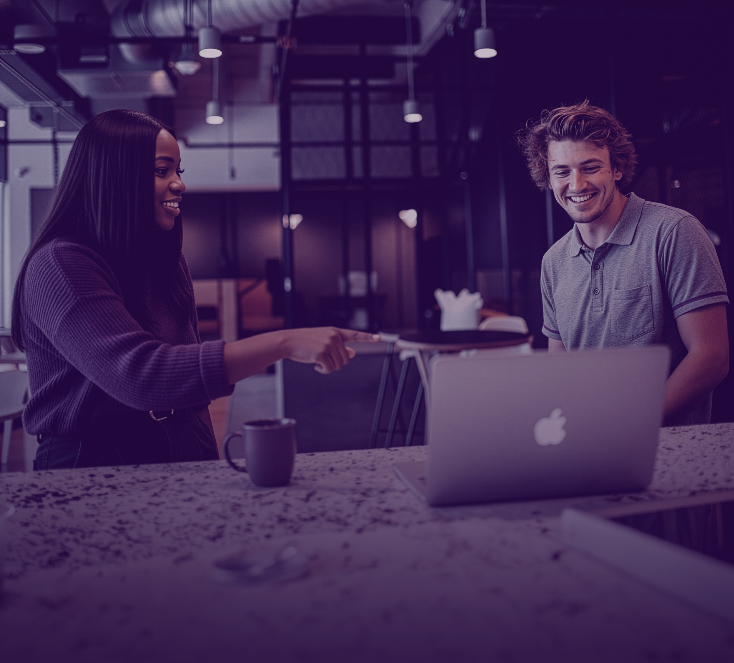 A woman pointing at a laptop screen while smiling, and a man sitting beside her also smiling in a modern office setting.