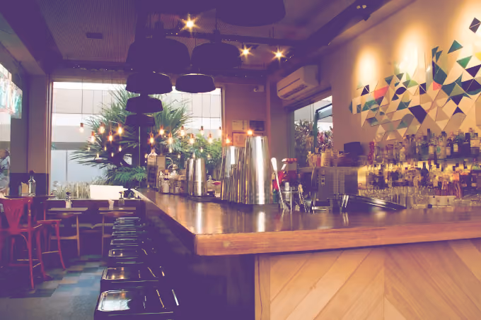 Modern bar interior with wooden counter, bar stools, hanging lights, and geometric wall art.